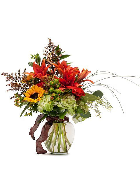 Autumn floral arrangement in a glass vase, featuring orange lilies, sunflower, and brown ribbon.