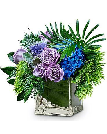 Floral arrangement with purple roses, blue hydrangea, and green foliage in a clear, square vase.