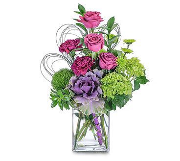 Floral arrangement in a clear square vase with purple roses, carnations, green hydrangeas, and ornamental kale.