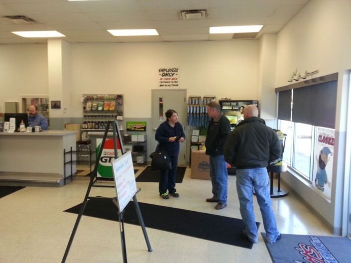 Three people stand in the lobby of an automotive shop, with a service desk in the background and signs on the walls.
