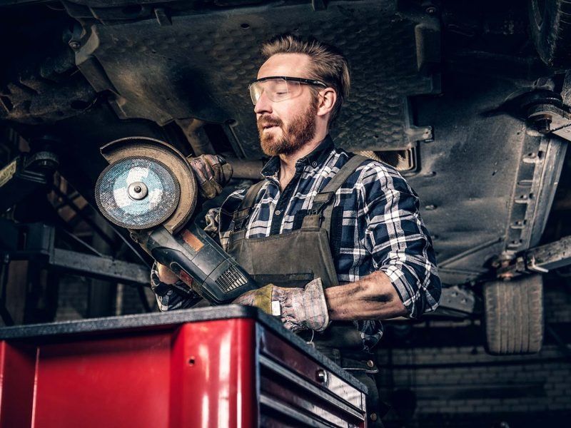 A mechanic in a plaid shirt and overalls wears safety glasses while holding an angle grinder in a car repair shop.