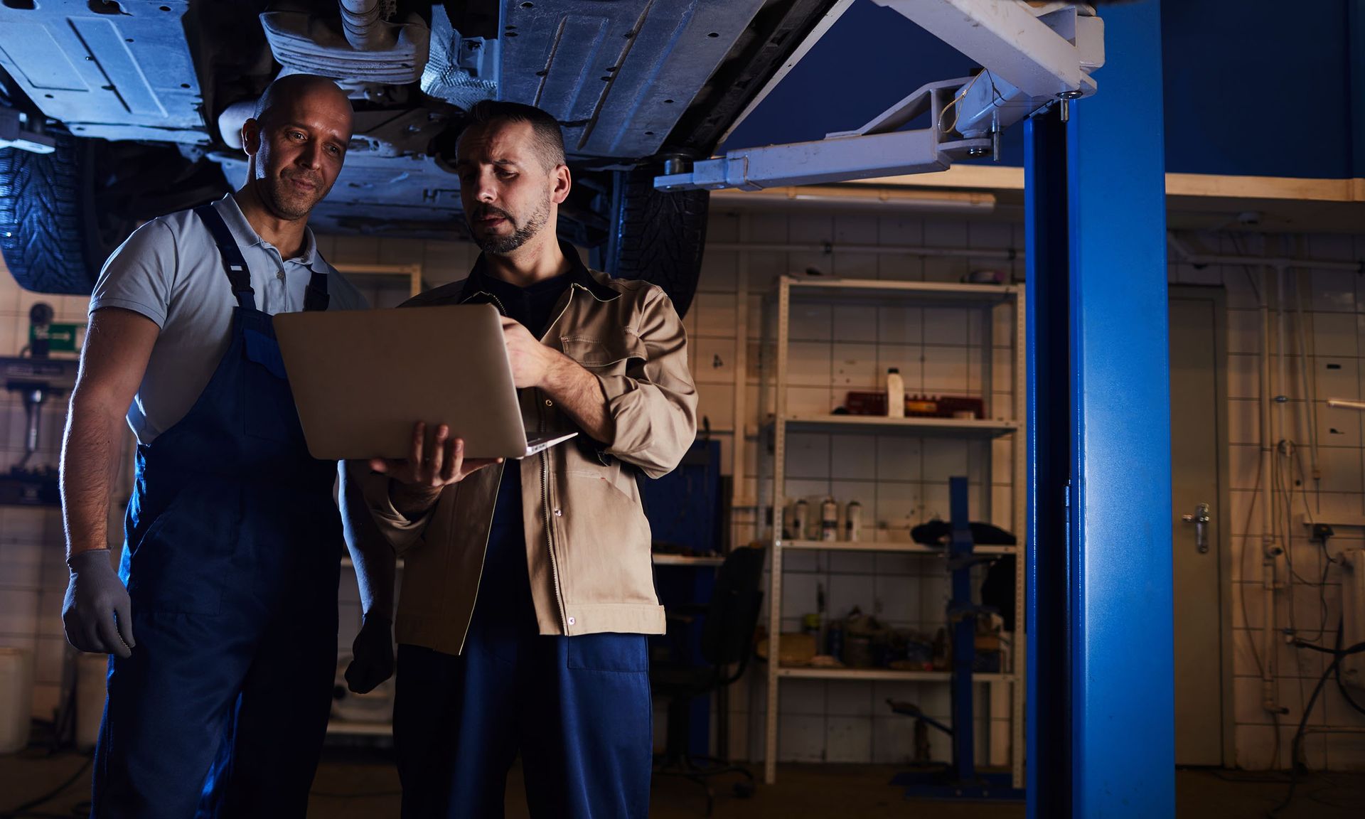 Two mechanics in a garage stand under a lifted car, reviewing information together on a laptop screen.