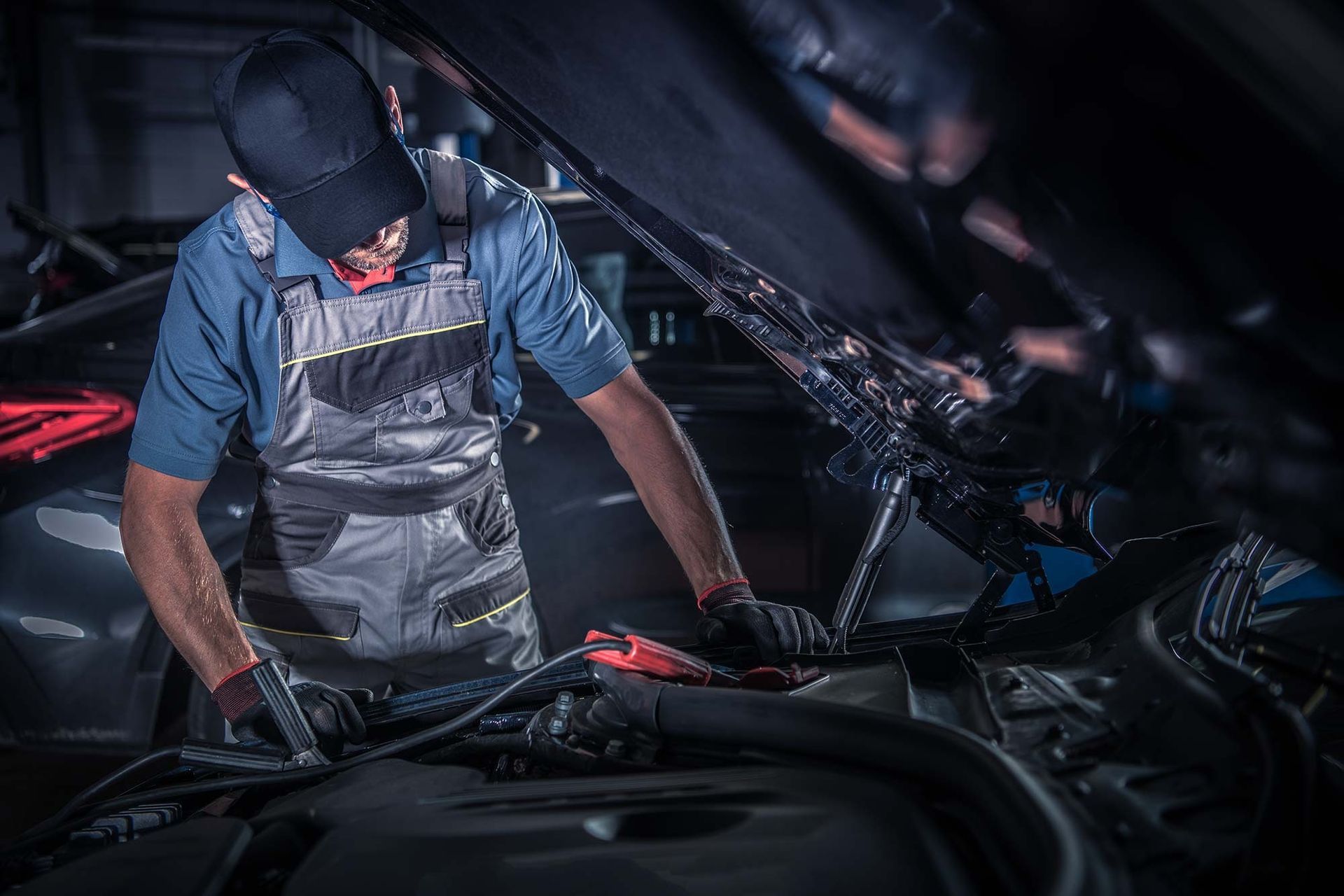 A mechanic in work overalls and a baseball cap repairs a car engine under the hood in a dimly lit garage.