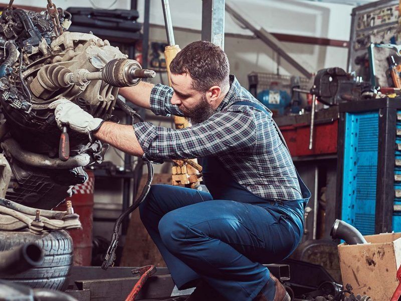 A focused mechanic in a plaid shirt and blue overalls works on a car engine in a cluttered garage.