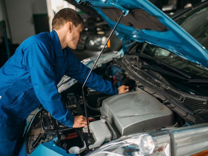 A mechanic in a blue uniform leans into the open hood of a car to inspect the engine inside a repair shop.