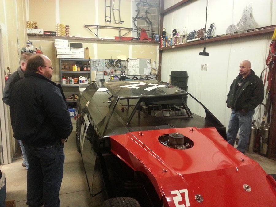 Three people stand in a garage discussing a red and black race car marked with the number 27.