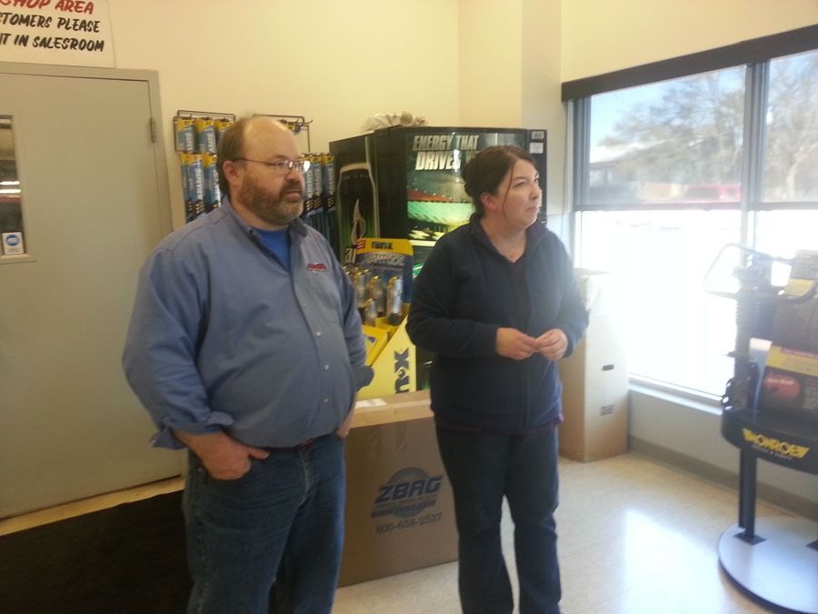 Two people stand in an office or retail space near a window, boxes, and a metal door.