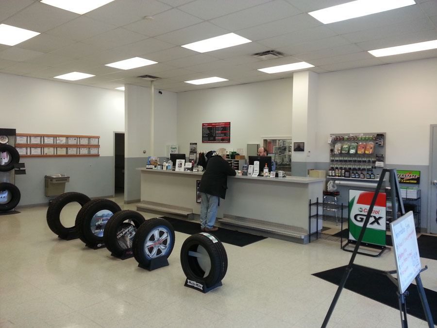 A customer stands at the service counter of an auto repair shop, with several tires displayed on the floor in the front.