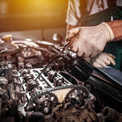 A mechanic in gloves pours oil from a small can onto the metal camshaft and gears of an exposed car engine.