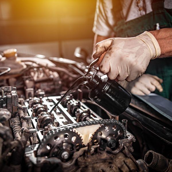 A mechanic in gloves pours oil from a small can onto the metal camshaft and gears of an exposed car engine.