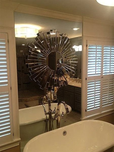 Bathroom mirror above a sink and bathtub, with white shutters and a sunburst wall decor reflection.