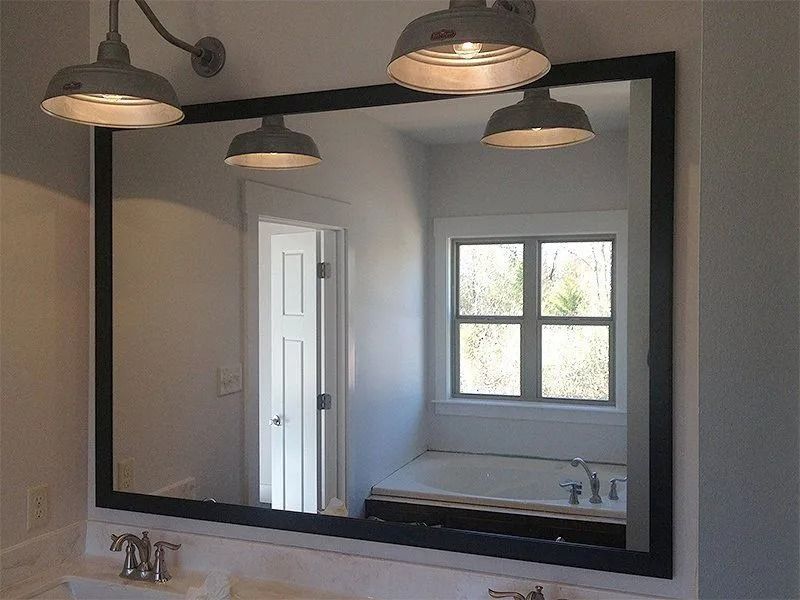 Bathroom mirror above sink reflecting a white door and window, with black frame and pendant lights overhead