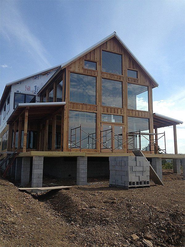 Partially built modern house with wood-framed windows and raised foundation on a dirt lot