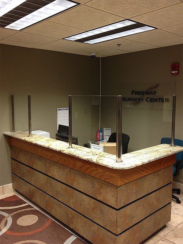 Empty hospital reception desk with beige counter, clear plexiglass barrier, and overhead fluorescent lights