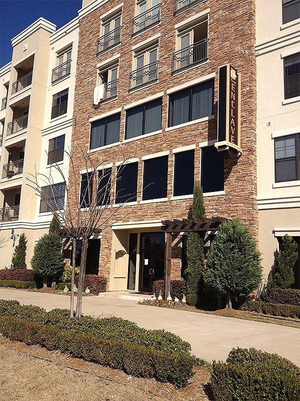 Brick apartment building with large windows and shrubs along the entrance walkway.