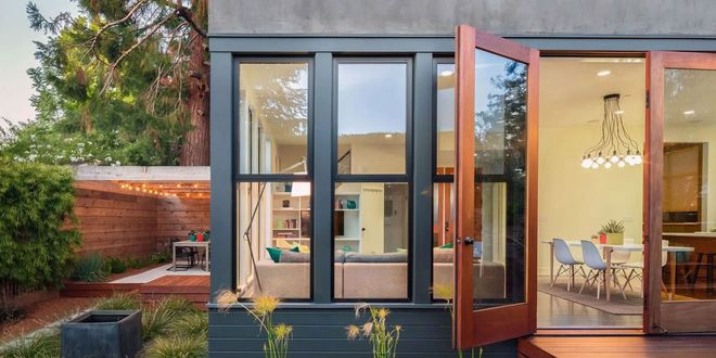 Modern glass house with open wooden door, lit dining area, and landscaped yard at dusk
