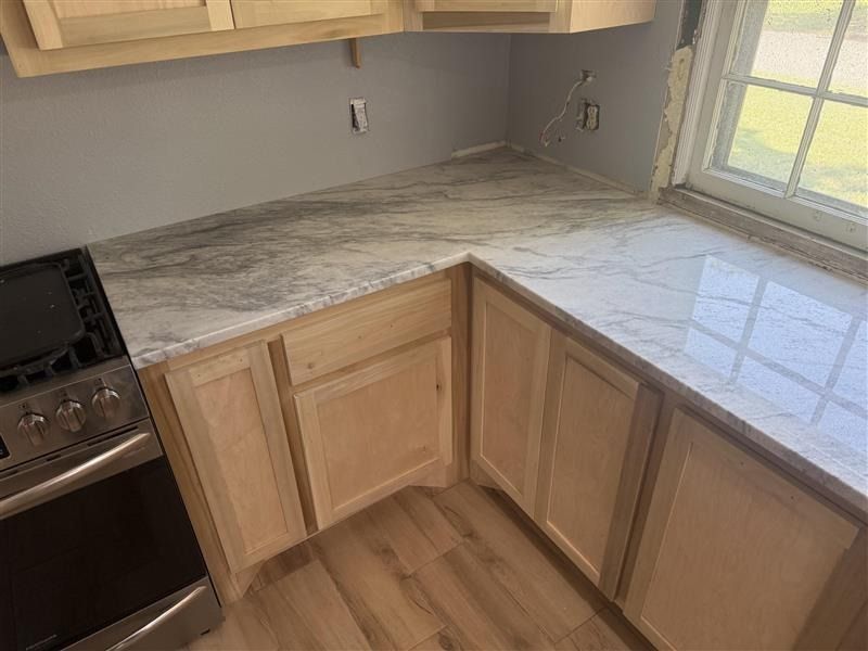 Empty kitchen corner with light wood cabinets, gray countertop, stove, and window.