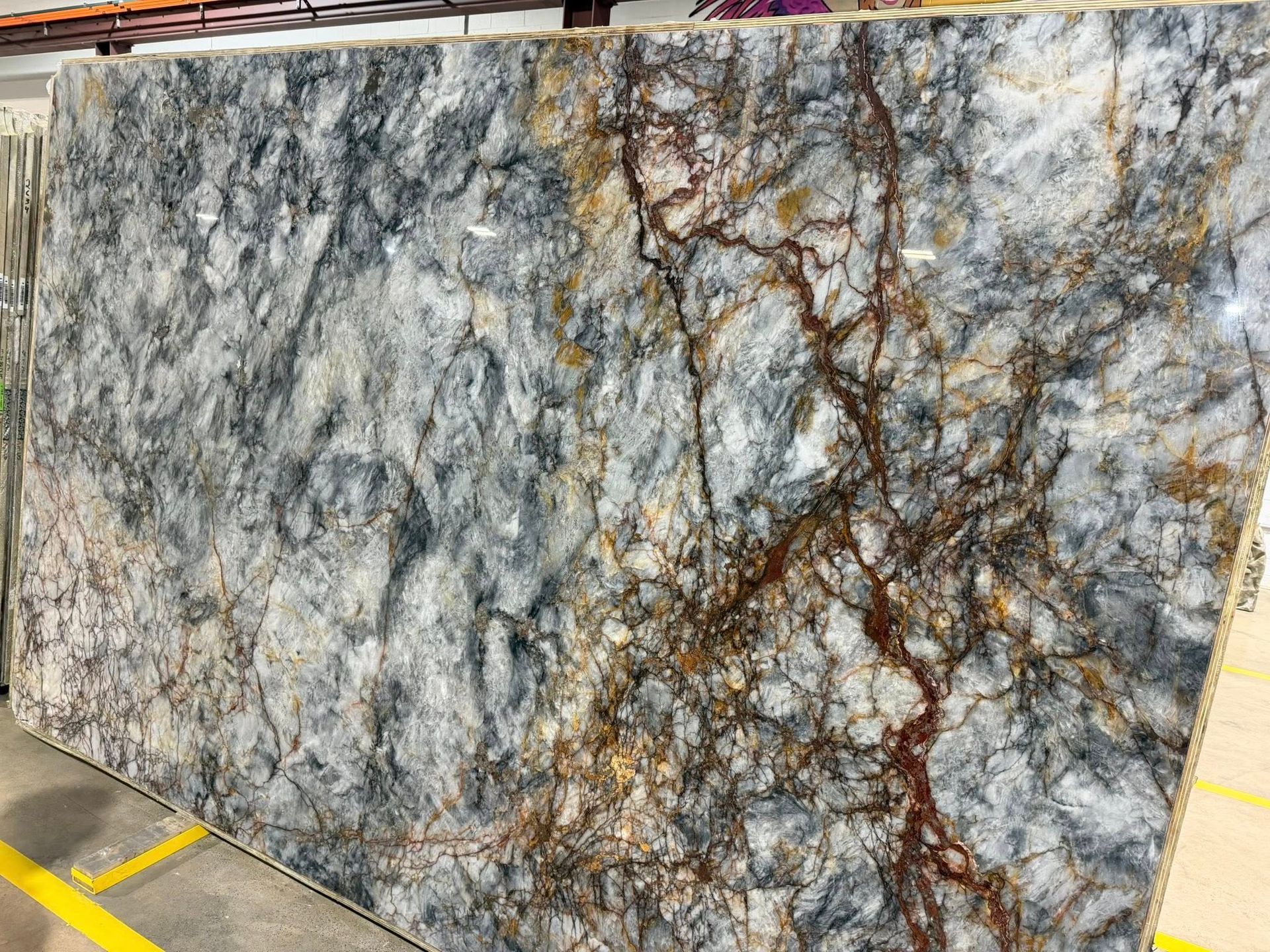 Large polished stone slab with gray, white, and brown veining in a warehouse setting