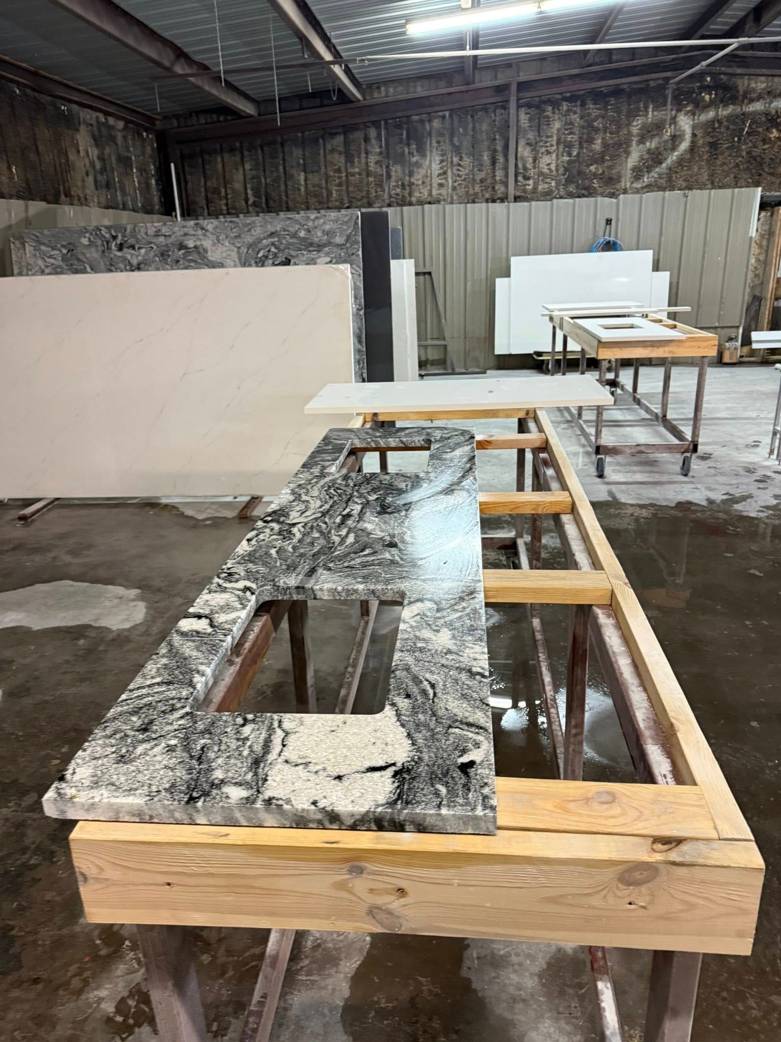 Stone countertop slab on a wooden frame in a workshop, with other work surfaces and materials nearby.