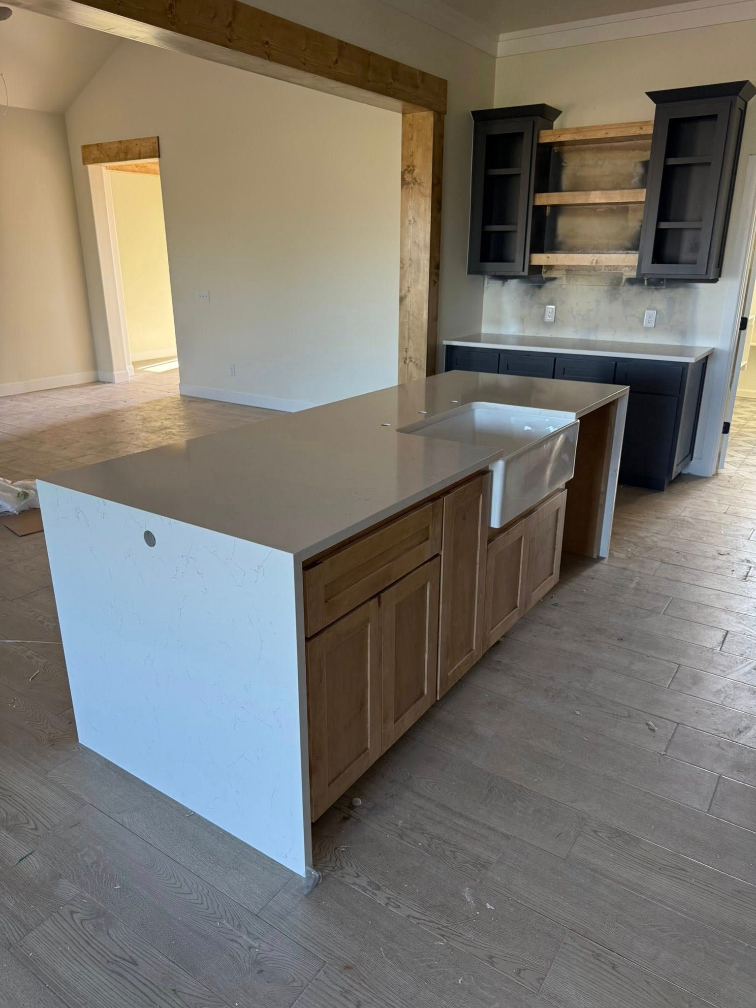 Empty kitchen with island, white countertops, wood cabinets, and dark upper cabinets against a neutral wall.