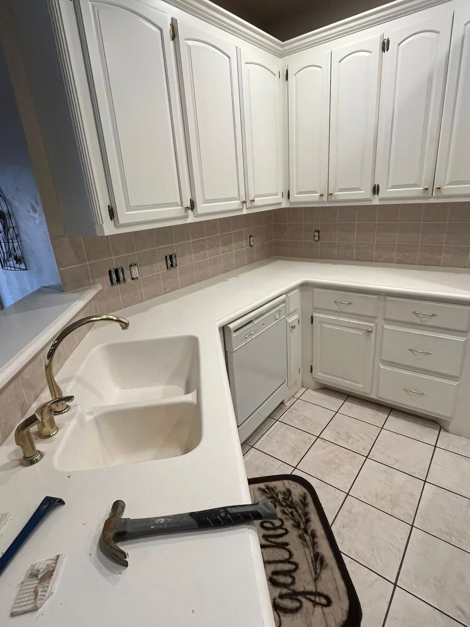White kitchen with tiled floor, corner cabinets, sink, and countertop with tools laid out during work.