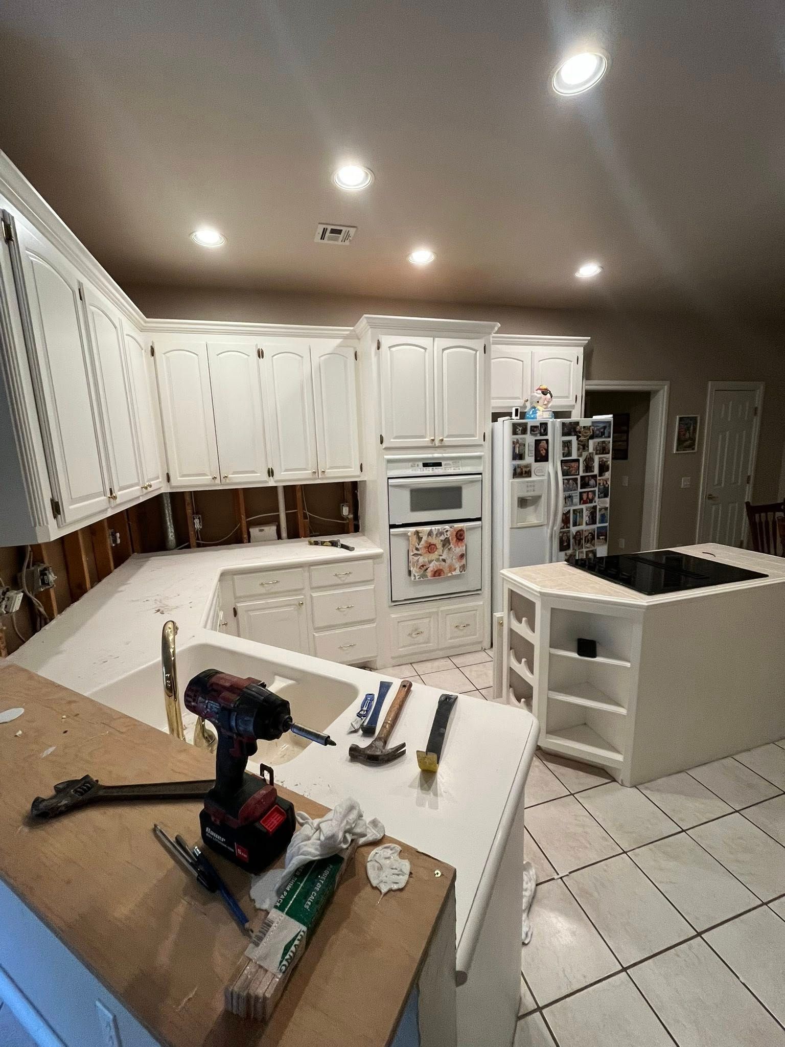 Bright white kitchen with tiled floor, island, refrigerator, and tools on the counter during a renovation.
