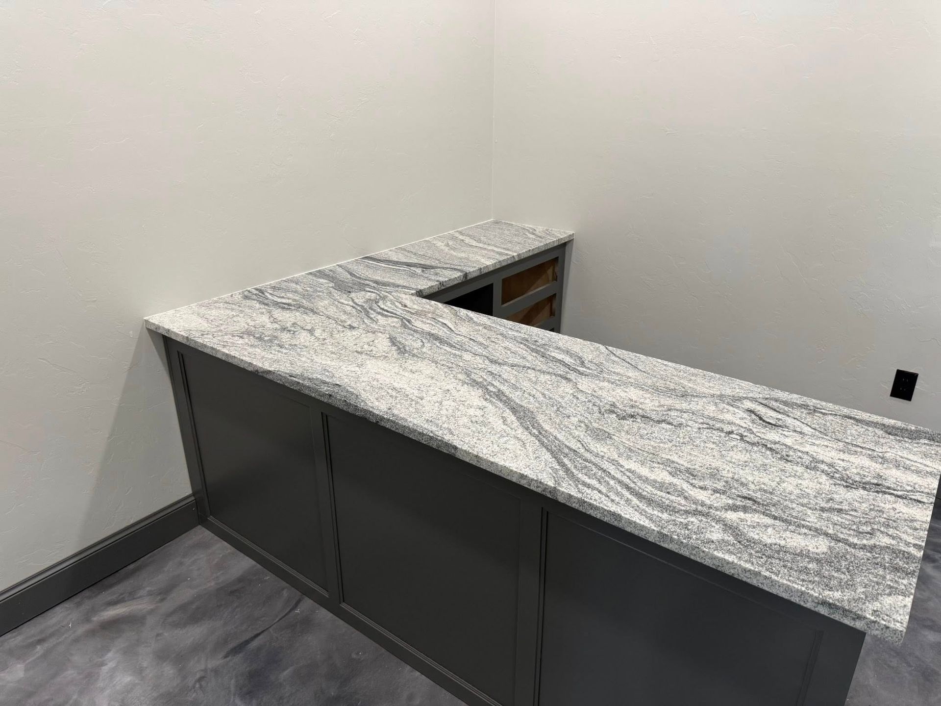 L-shaped gray stone countertop on dark cabinets in an unfinished room with white walls and gray floor