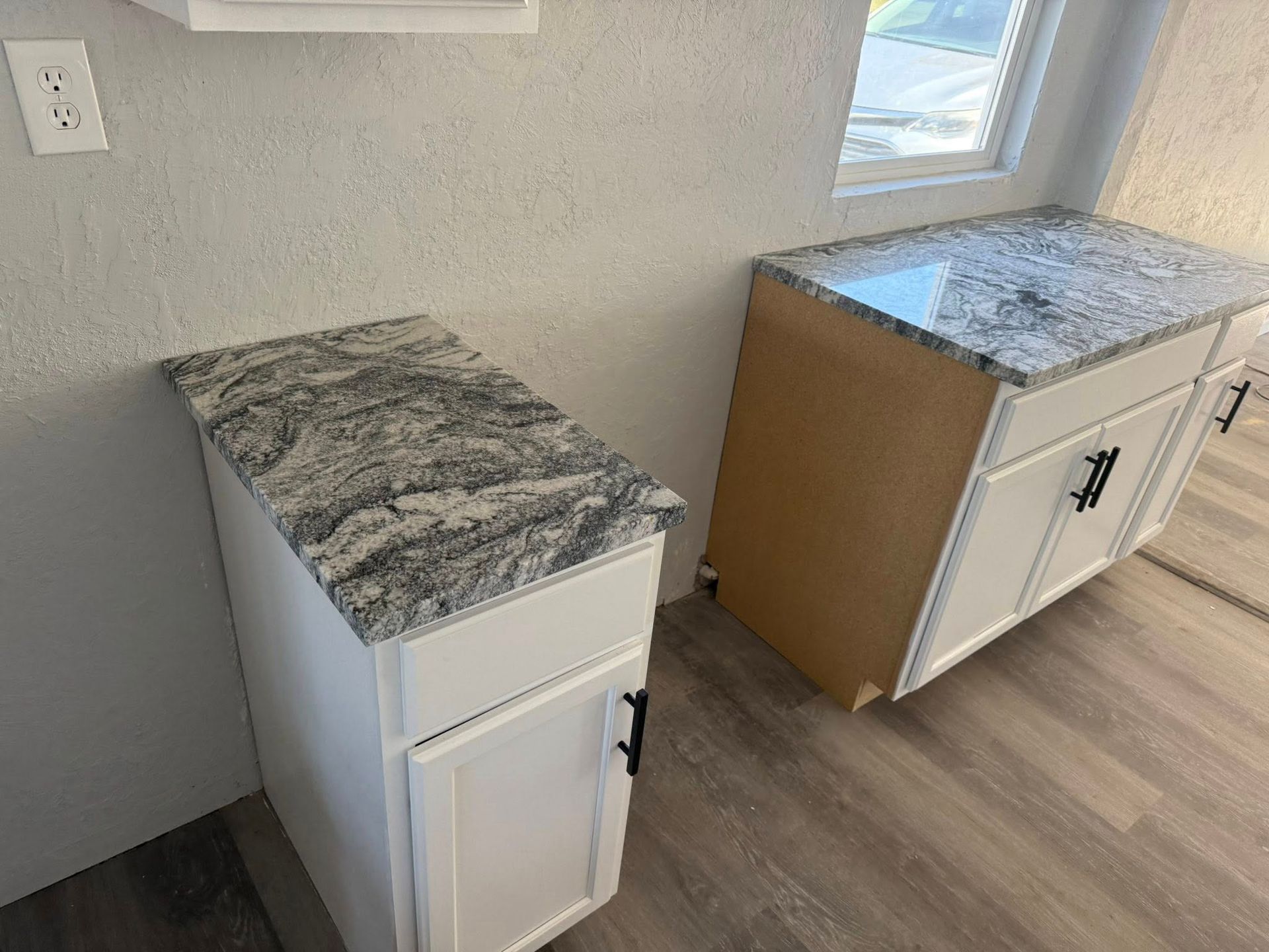 Two small white cabinets with marble-look tops against a wall near a window in a bright room