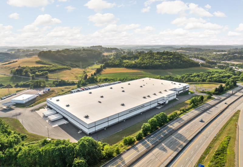 Aerial view of a large warehouse with a flat roof, next to a highway, in a rural landscape.