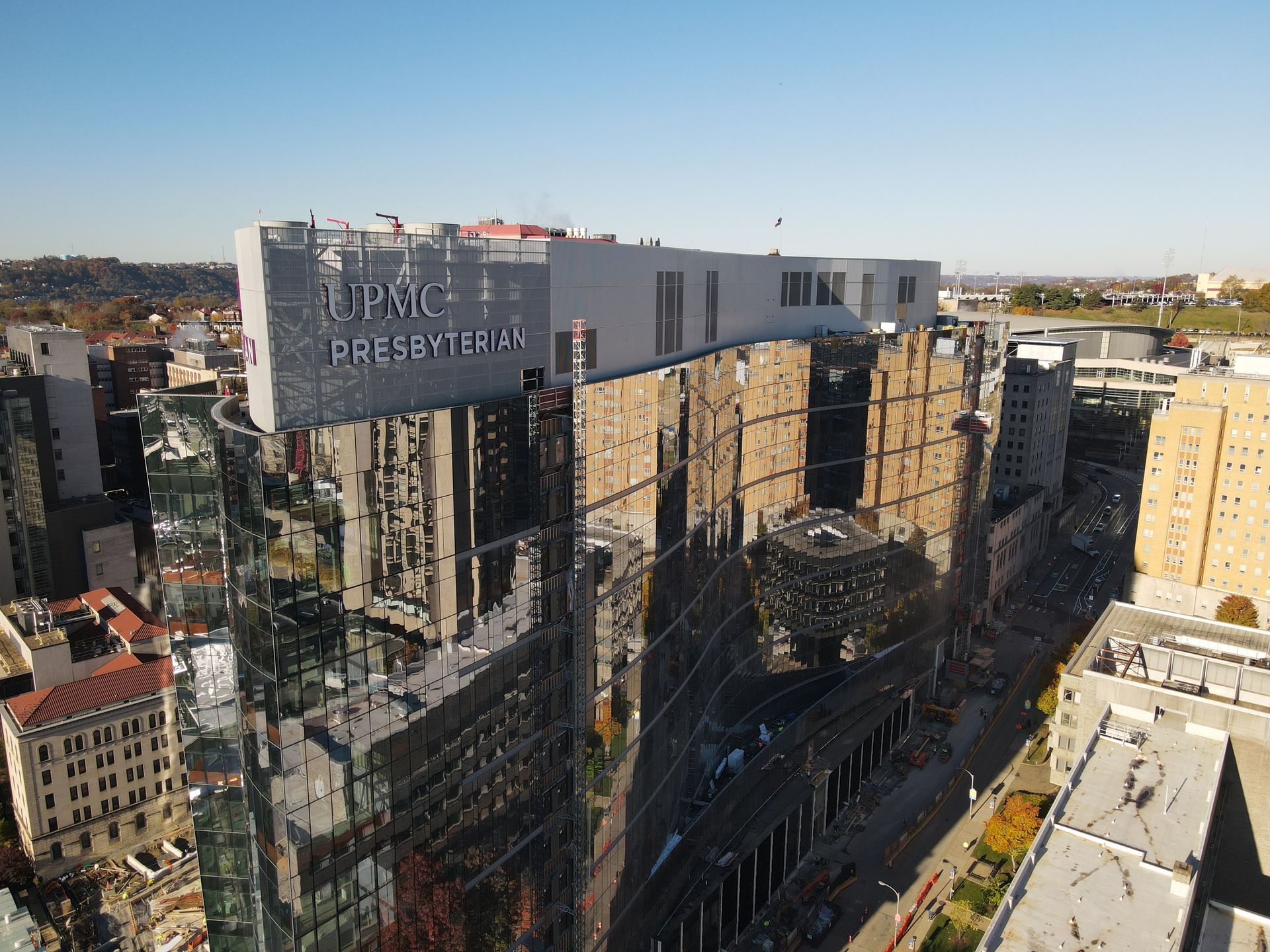Reflective glass skyscraper, “UPMC PRESBYTERIAN” sign on top, sunny urban setting.