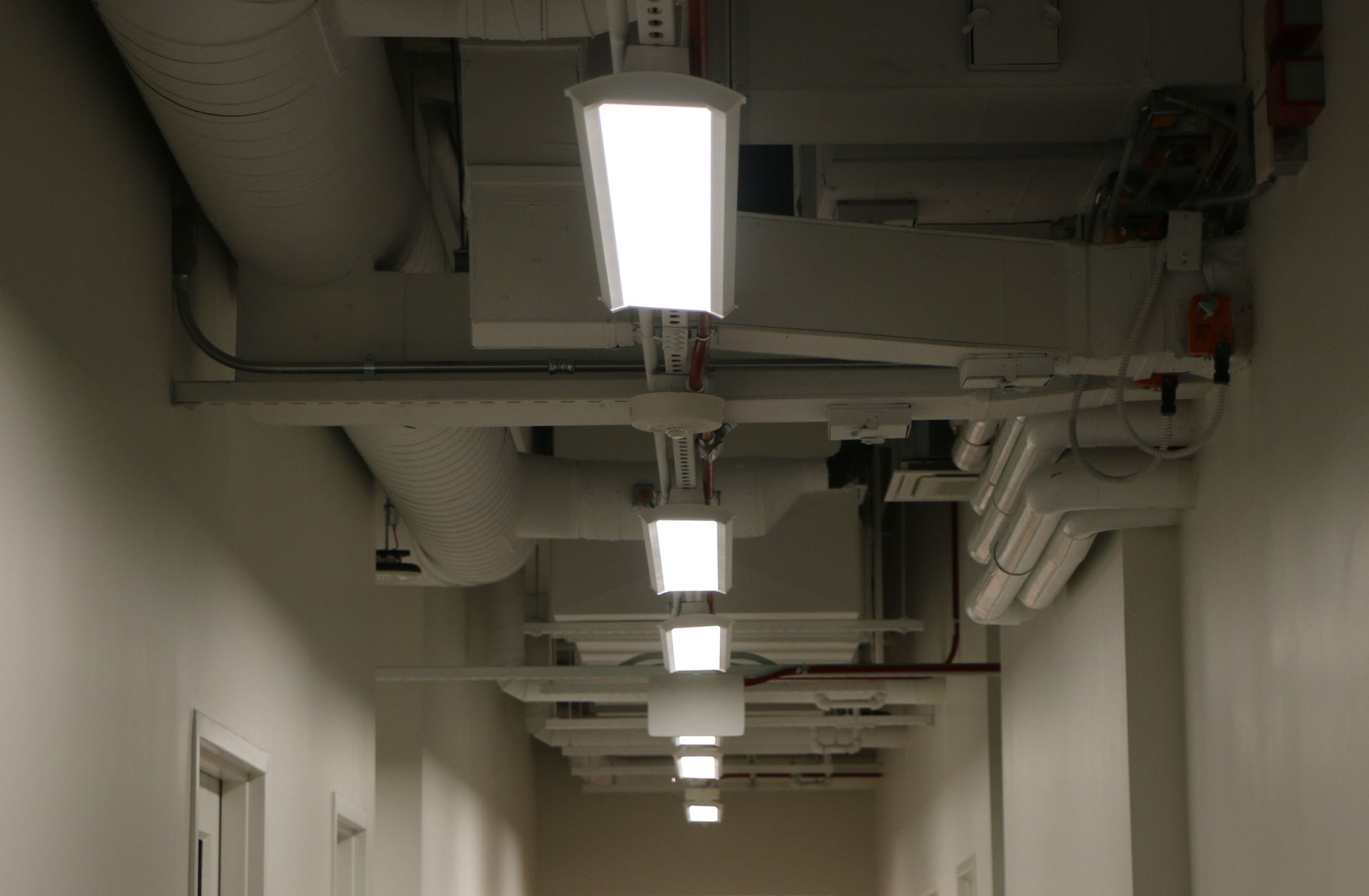 A hallway with bright rectangular lights hanging from the ceiling, along with pipes and ducts.