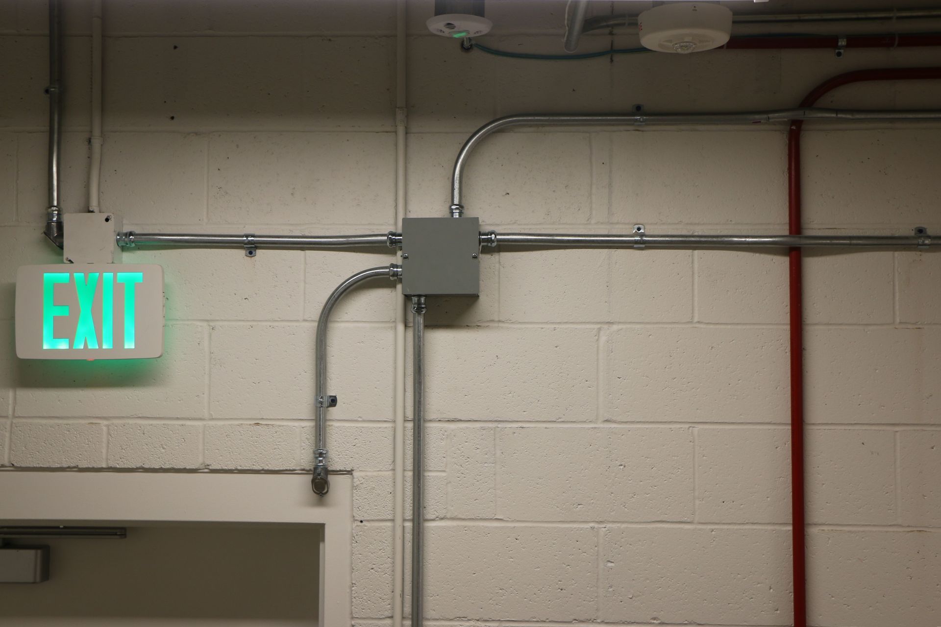 Green EXIT sign on white cinder block wall with electrical conduit.