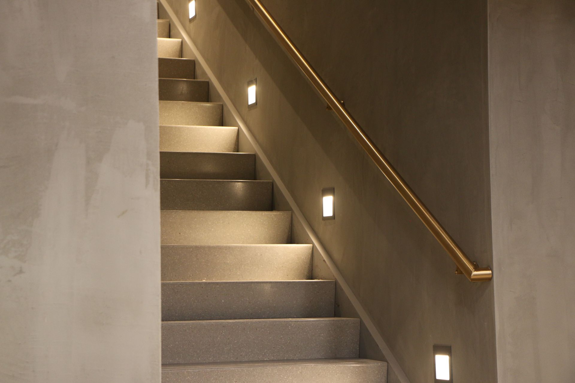 Staircase with built-in lights, and a gold-colored handrail, lit from below. Gray walls and steps.