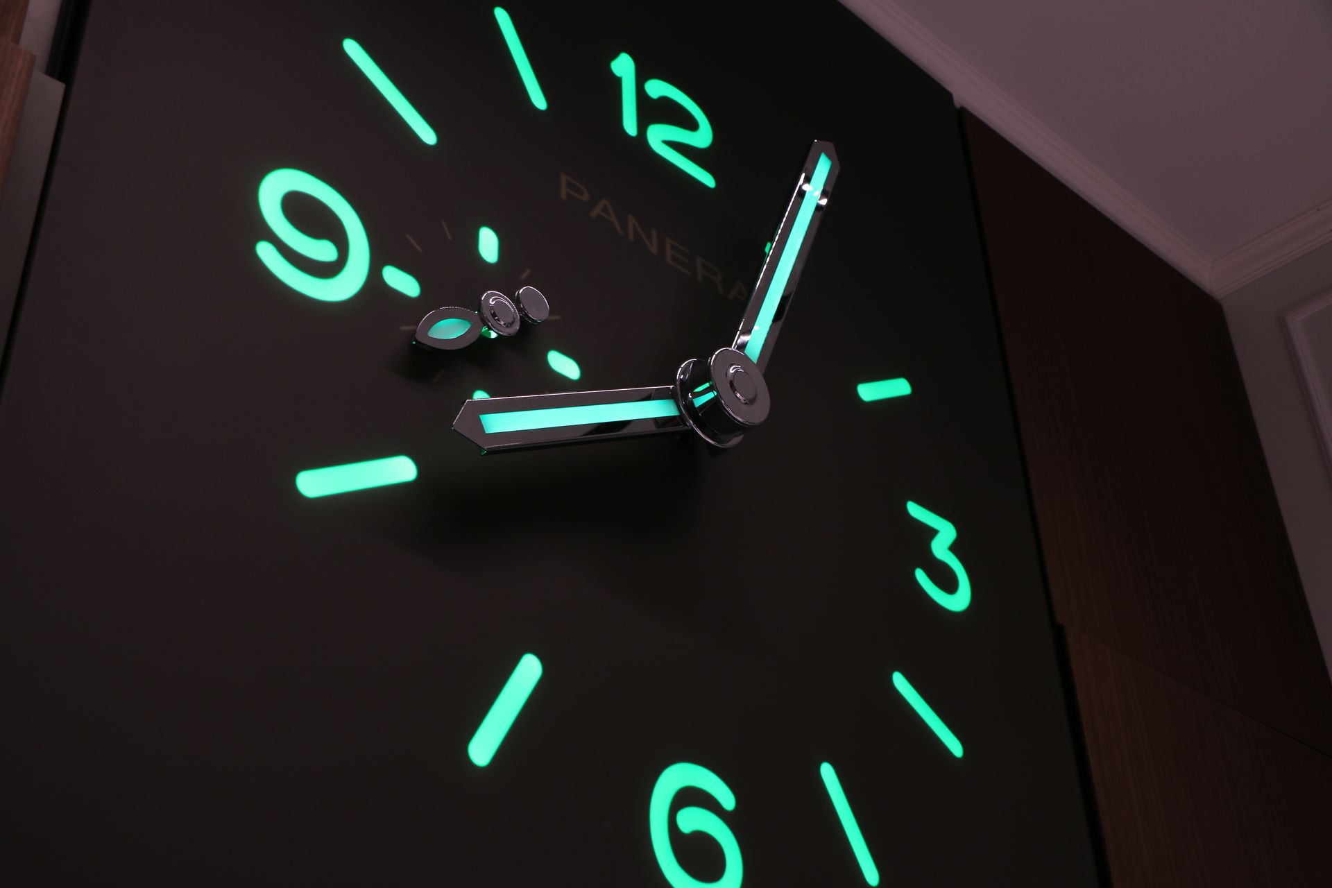 Luminous black wall clock with bright green numerals and hands; brand name "PANERAI" is visible.