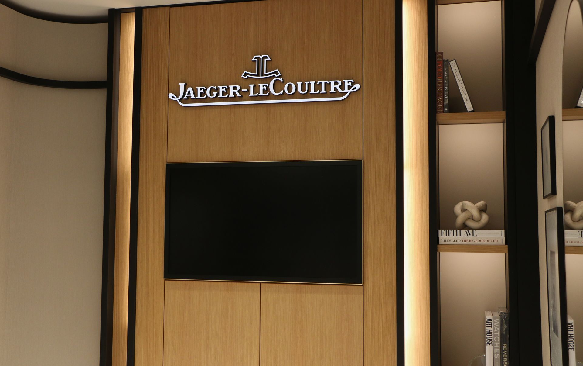 Jaeger-LeCoultre logo above a black screen, mounted on a wooden wall. Bookshelves to the right.