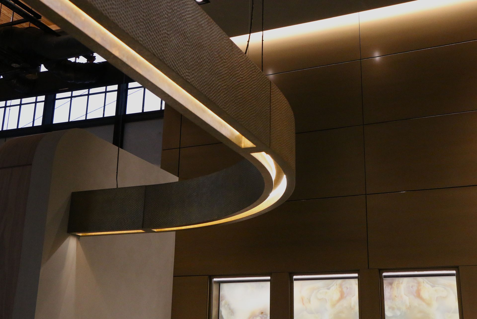 Curved, illuminated ceiling fixture in an interior space with a wall and windows.