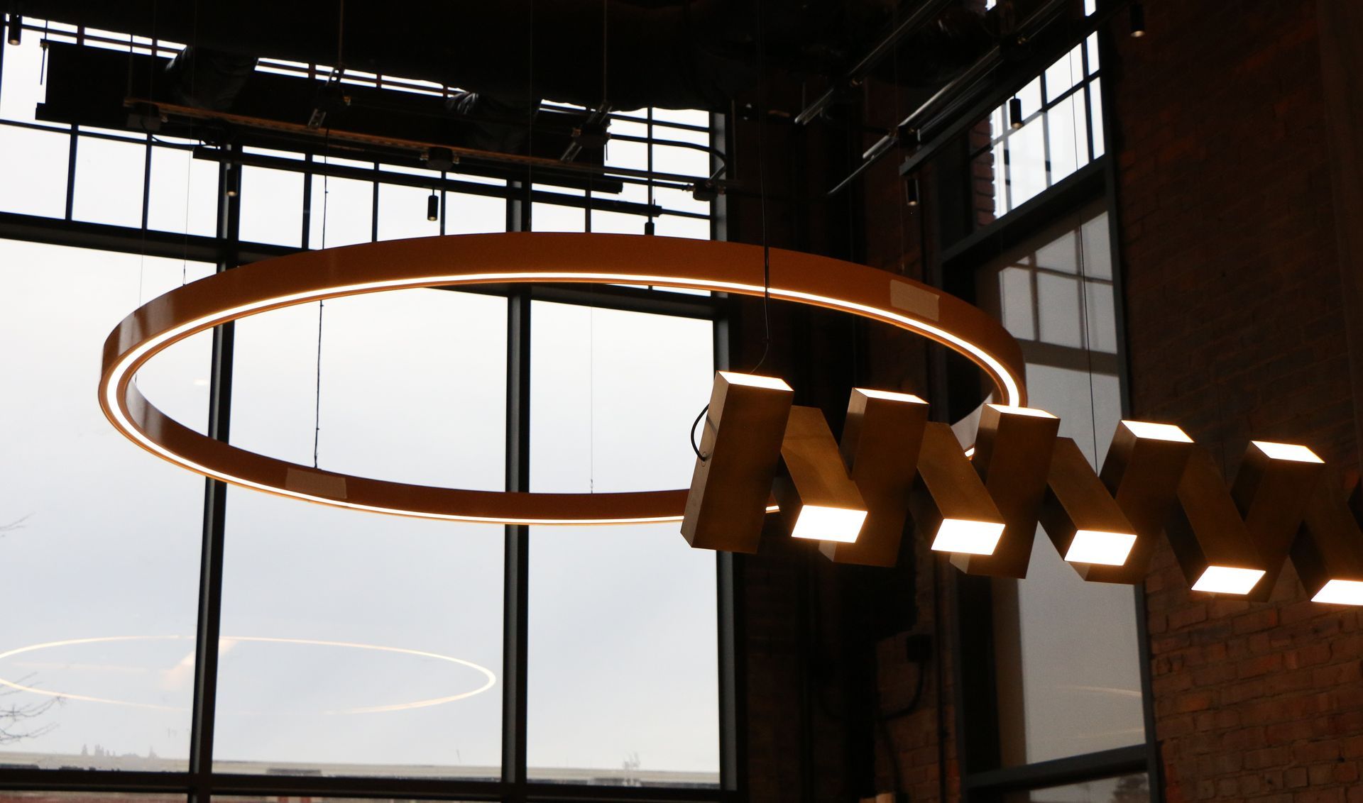 Large circular light fixture and geometric pendant lights in a high-ceilinged room with large windows.