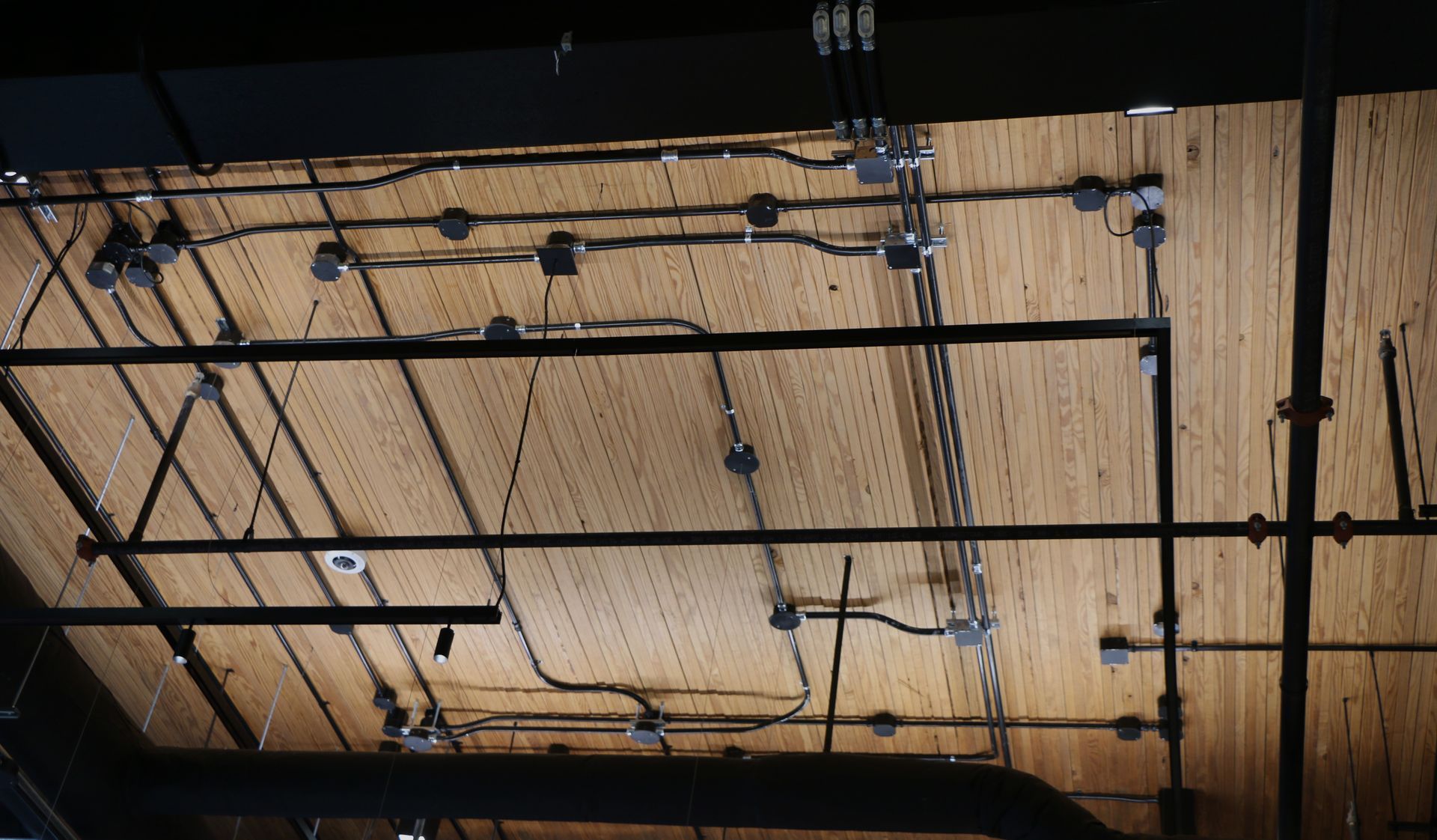 Ceiling with exposed wiring, pipes, and electrical boxes against a wood plank background.