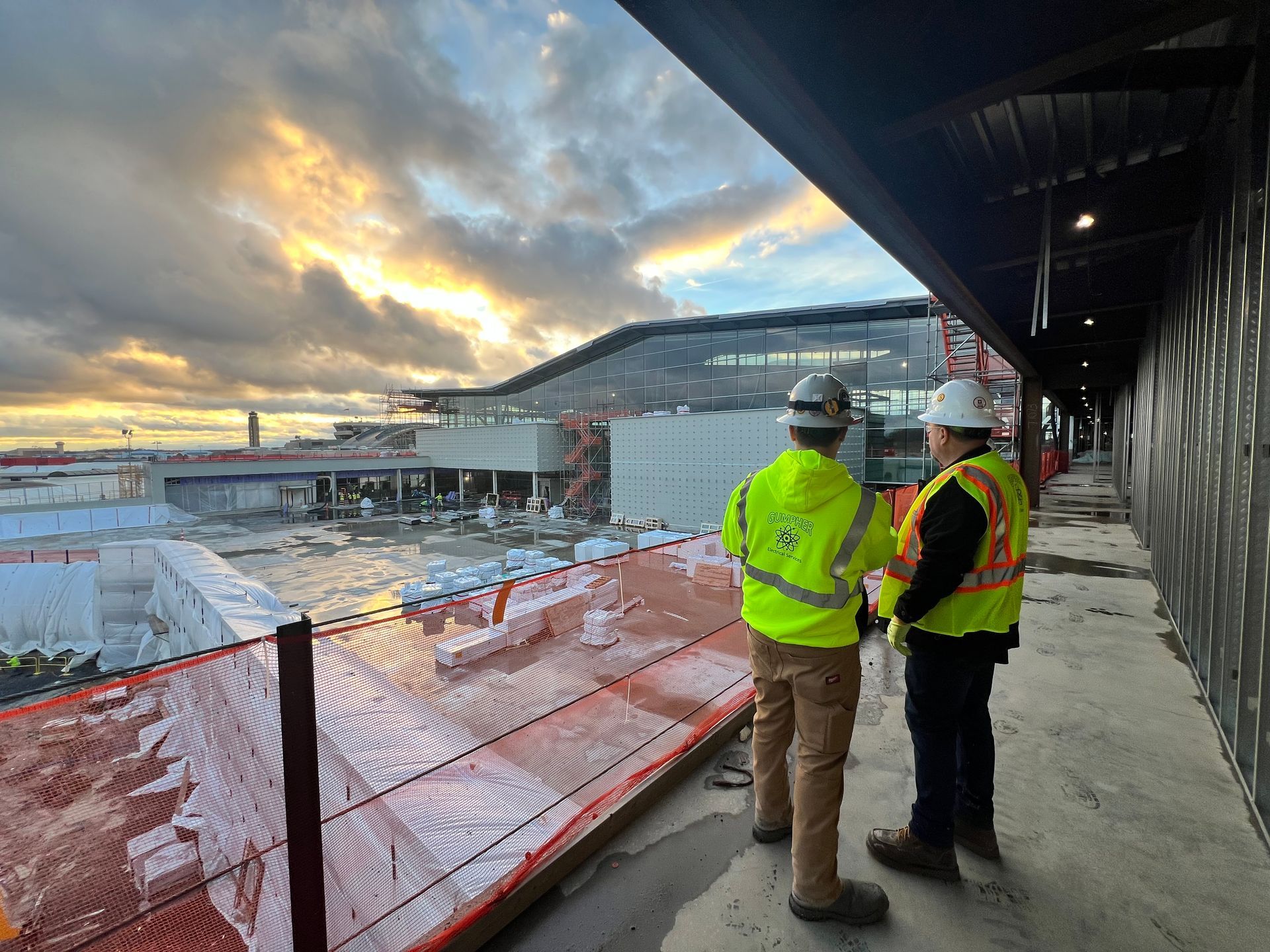Two construction workers in safety vests and hard hats surveying a building site at dusk.