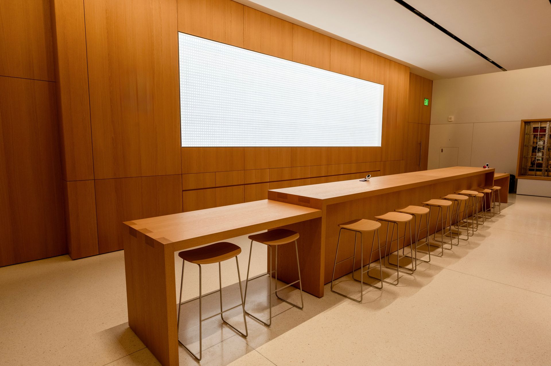 A modern room with wood-paneled walls, a large screen, long tables, and stools.