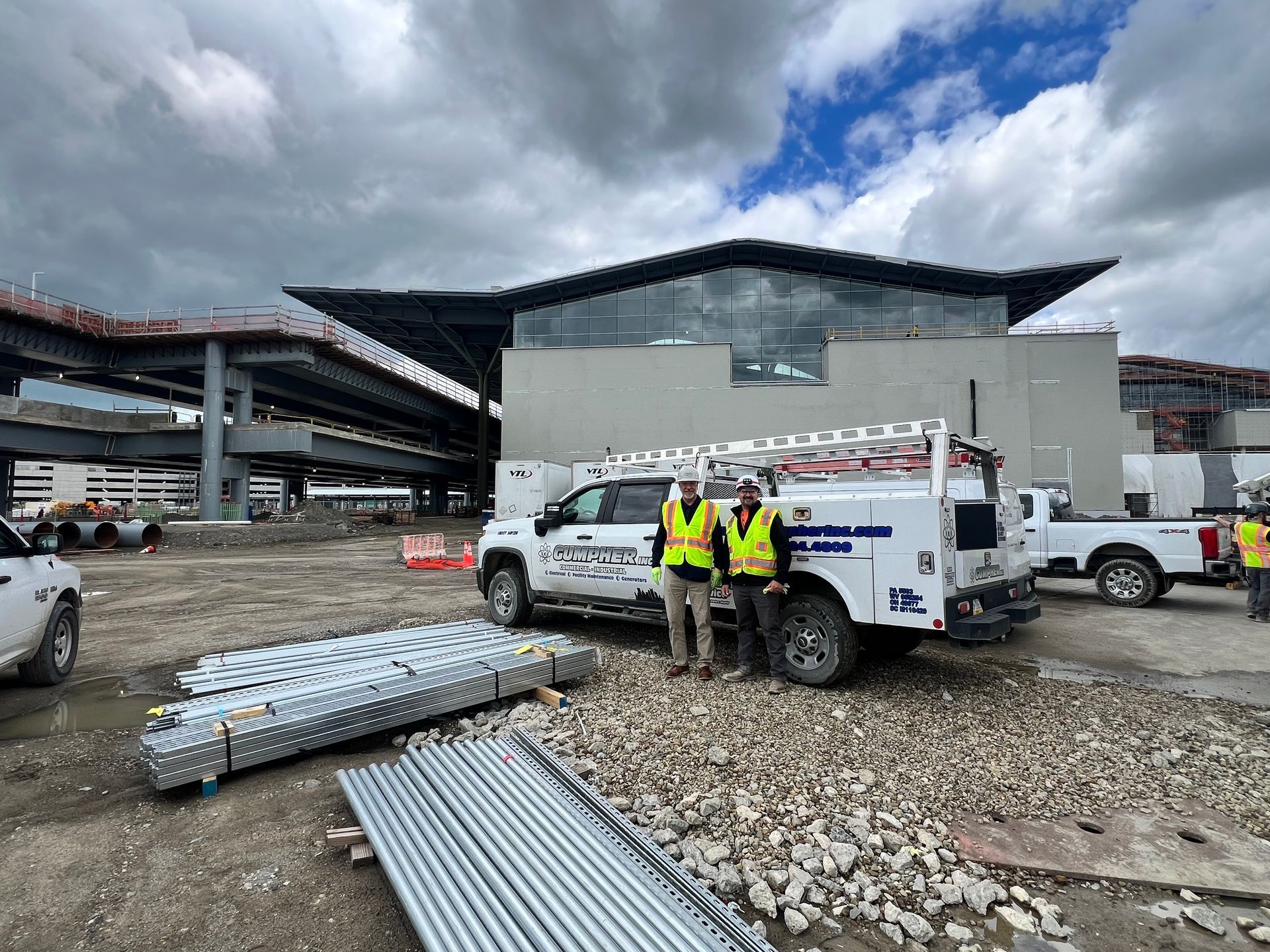 Three people in safety vests pose near a truck in front of a building under construction.