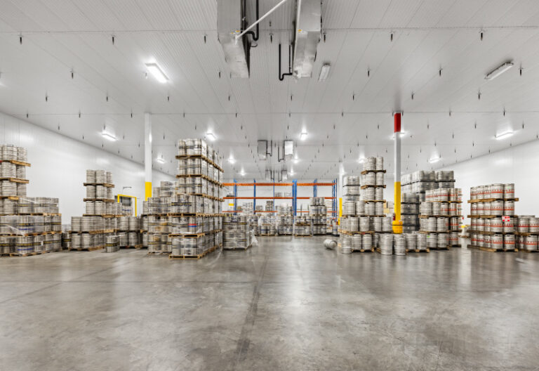 Warehouse interior with stacked pallets of boxes and barrels under bright lighting.