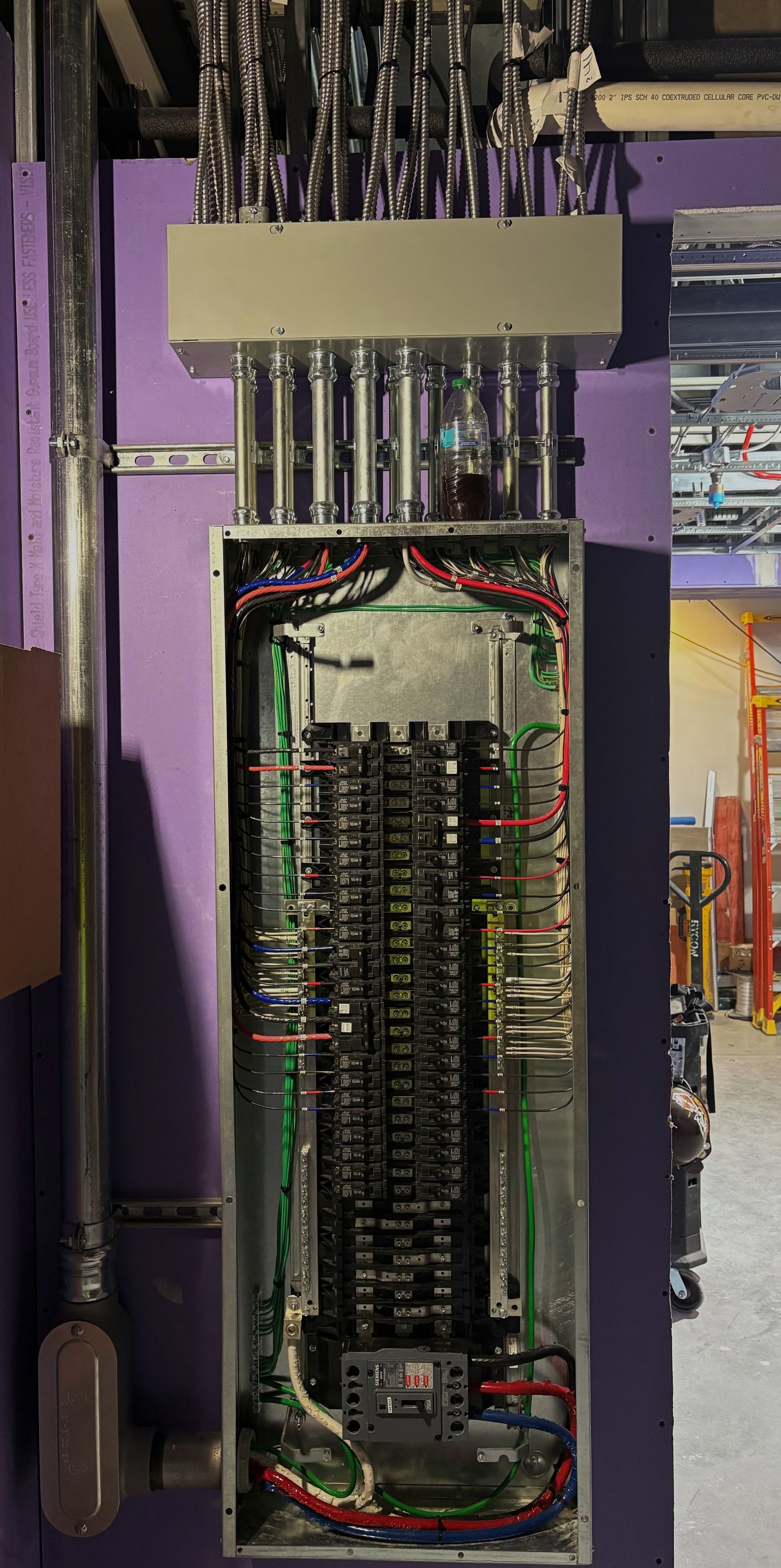 Electrical panel with many wires and conduits against a purple wall.