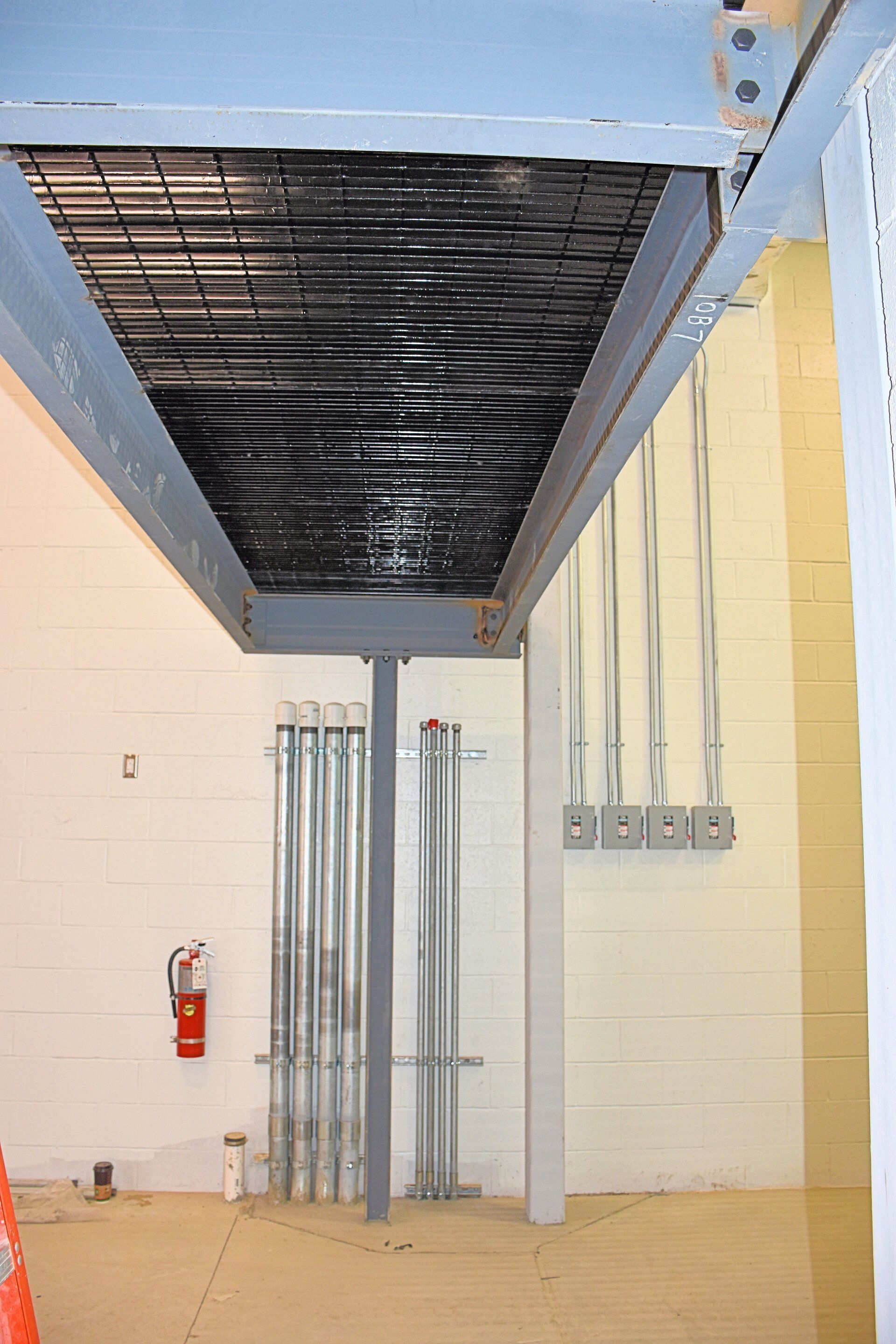 Metal grid ceiling with black covering, supported by a gray beam and pillar, over a mostly empty room with electrical conduits on the wall.