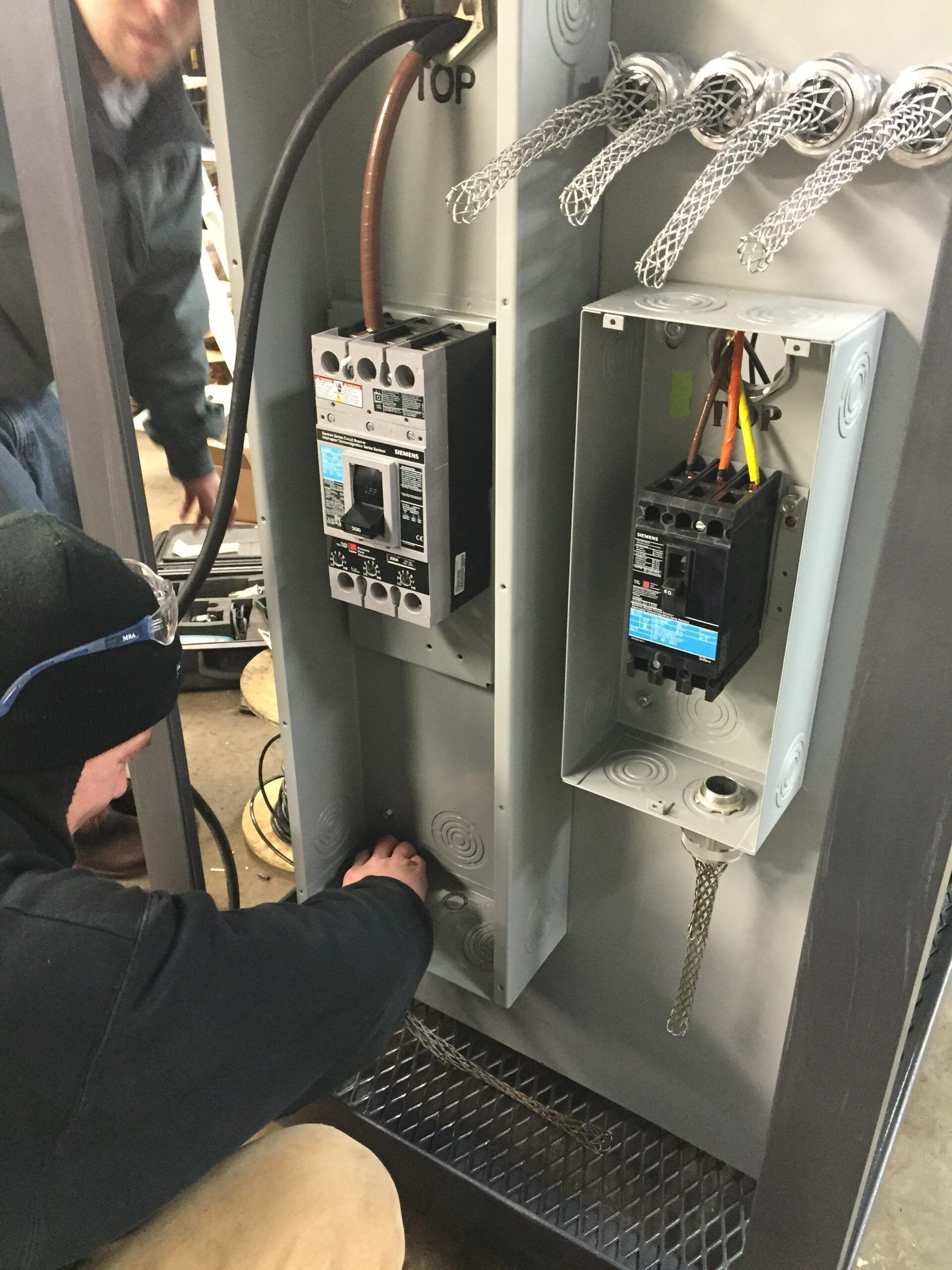 Person working on electrical panel with wires and circuit breakers. Another person watches.