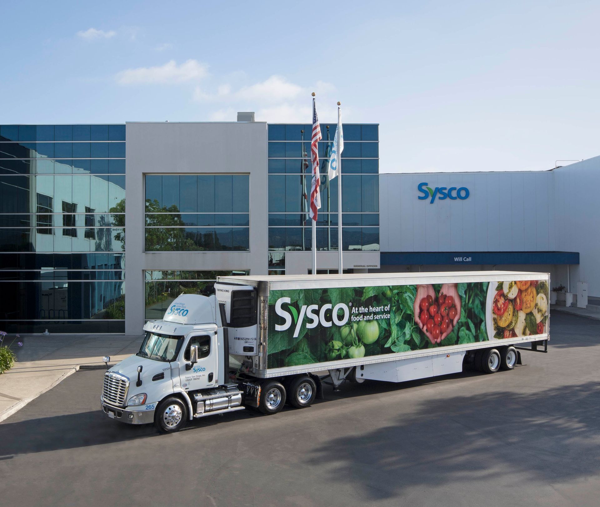 Sysco truck parked in front of a Sysco warehouse building with windows, under a bright sky.