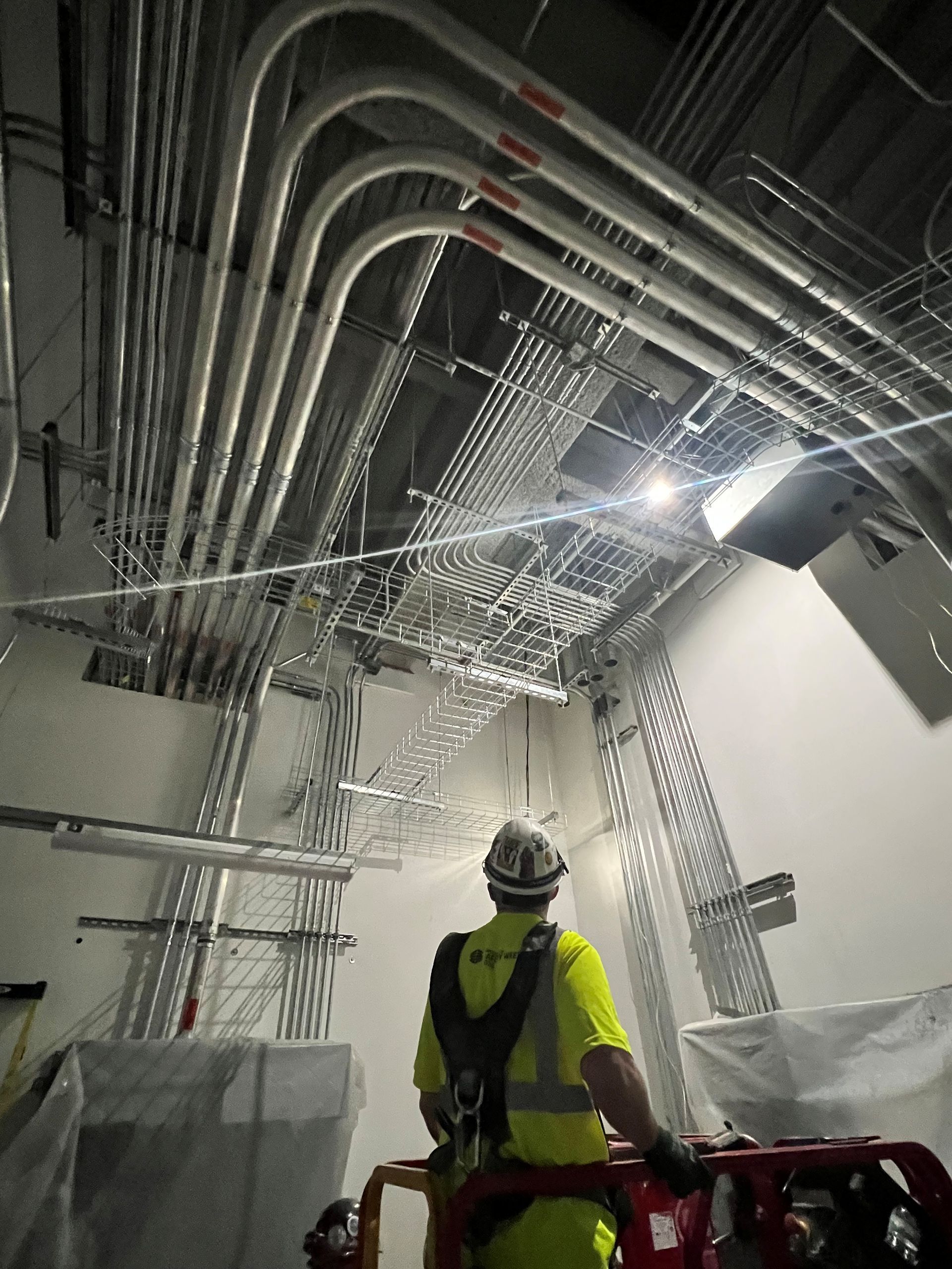 Worker in safety vest on lift, looking up at electrical conduit and cable trays installed on a ceiling.