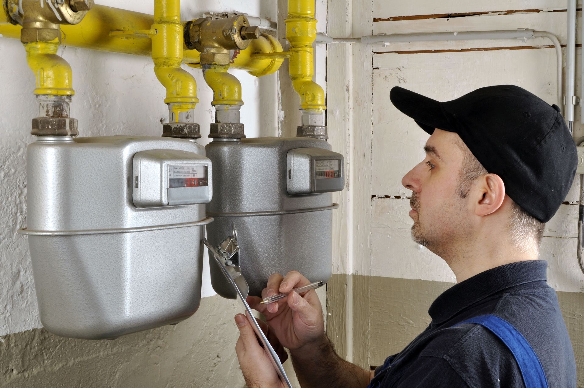 A person in a black cap inspects a gas meter with a wrench, indoors, near yellow pipes.