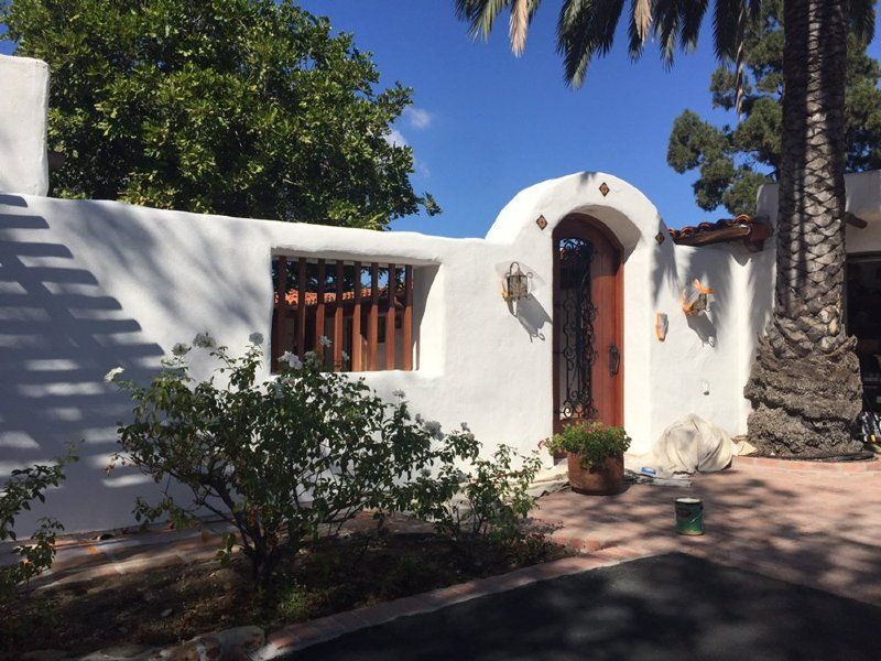 White stucco wall with arched wooden door and window, a small rose bush, and a palm tree against a sunny blue sky.