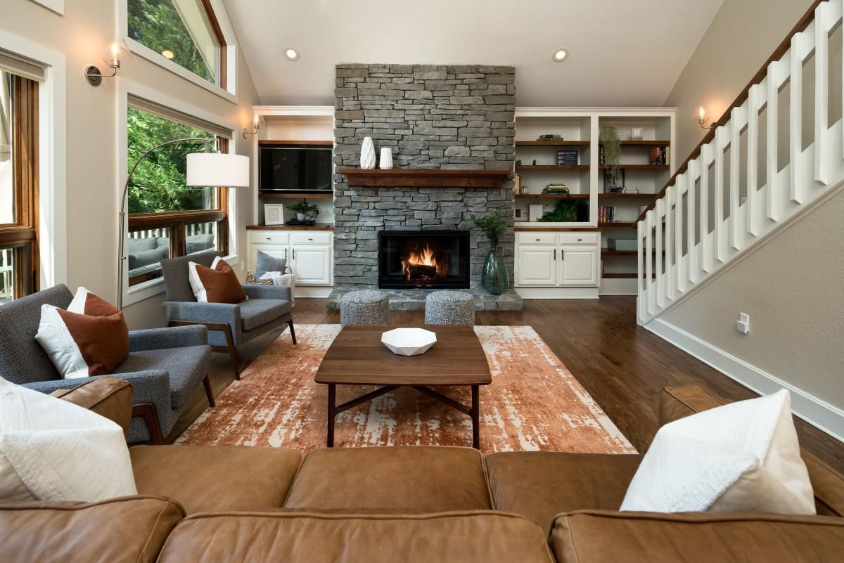 Cozy living room with stone fireplace, built-in shelves, and leather couch. Neutral tones with orange rug and pillows.